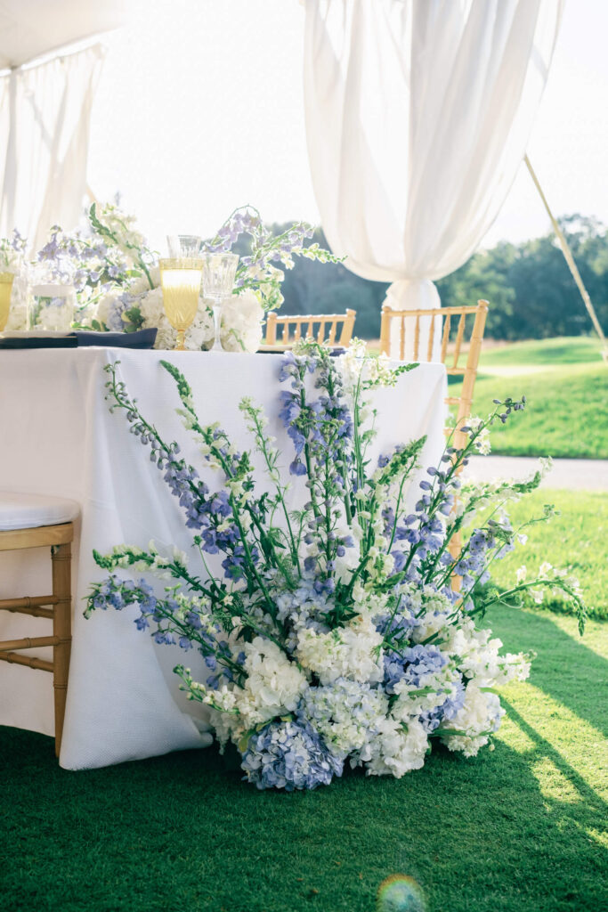 Close-up of cascading blue delphiniums and white hydrangeas spilling from the corner of a reception table under a tented wedding setup