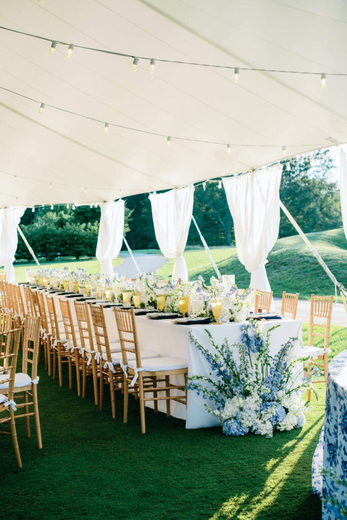 Wide view of the long reception table under an open-air tent with white draping and tall floral arrangements