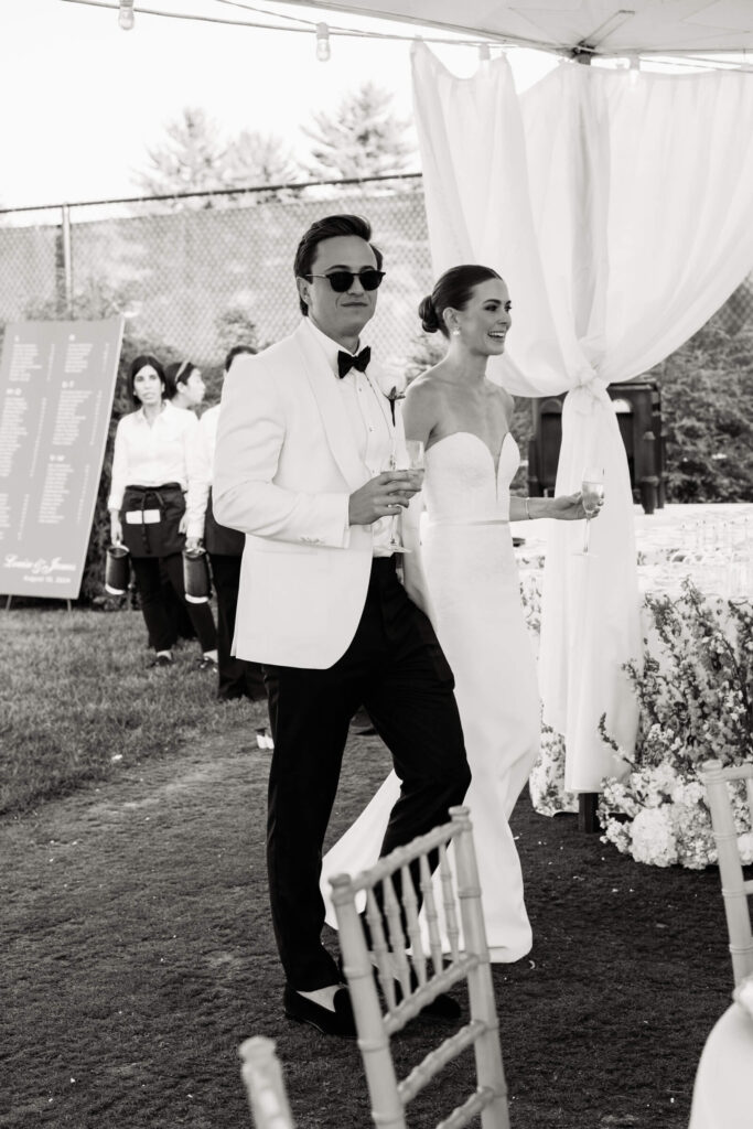 The bride and groom enter the reception tent smiling and holding champagne, surrounded by soft draping and floral arrangements