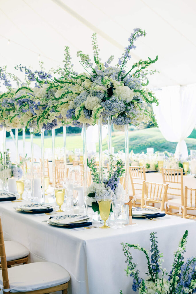 Head table set with tall glass vases overflowing with blue and ivory flowers and golden chairs lined on each side