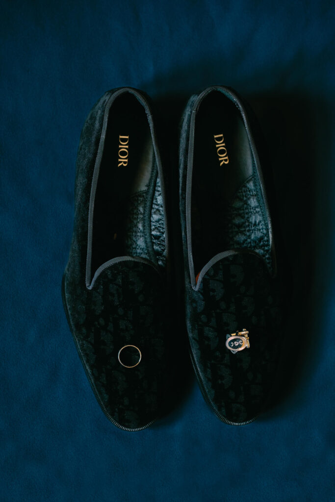 Flat lay of groom’s black Dior loafers with his gold wedding band and monogrammed cufflinks placed on a rich navy backdrop