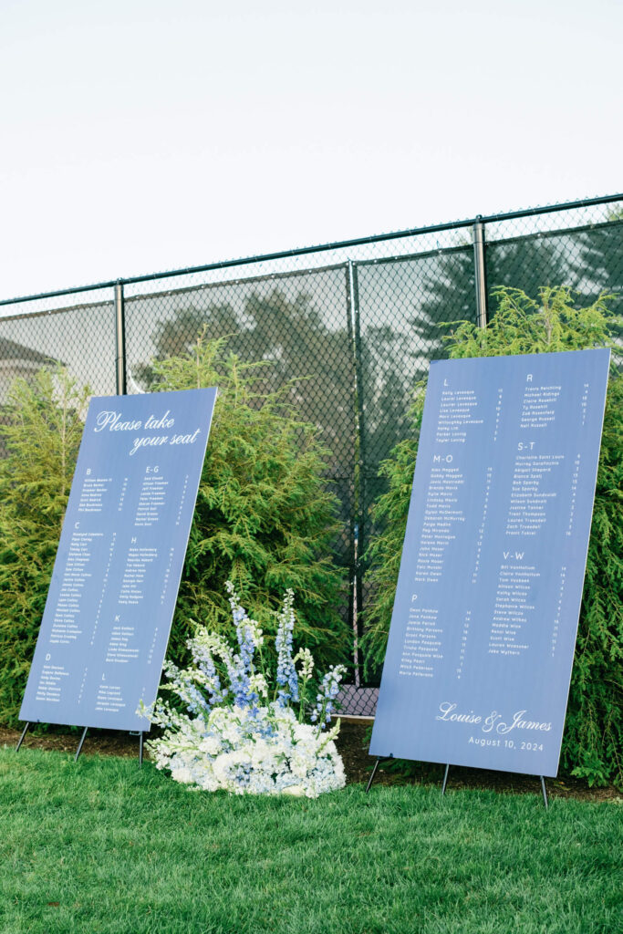 Elegant blue seating chart panels with white calligraphy and floral arrangements at the base, set against manicured hedges at a New York wedding celebration.