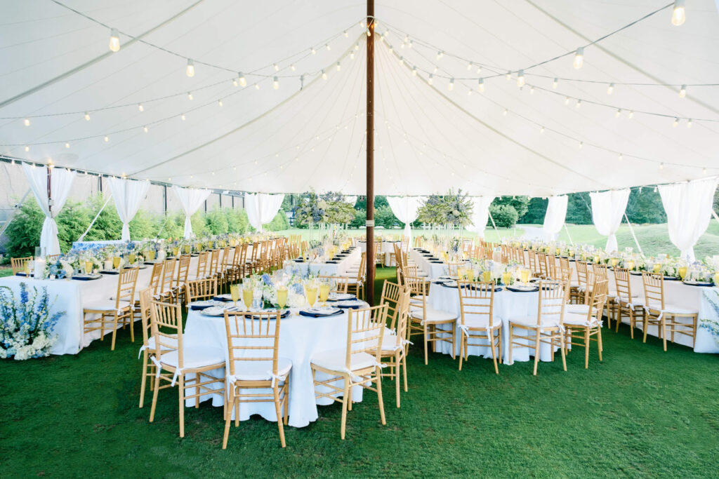 Wide-angle view of a sailcloth tent reception setup with glowing bistro lights, long and round tables, and lush garden-inspired floral centerpieces in cream and periwinkle.