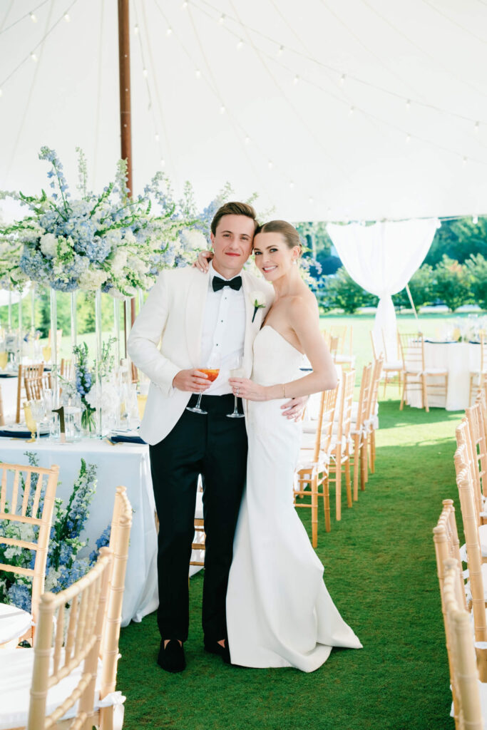 Bride and groom pose arm-in-arm inside their sailcloth tent reception, both holding drinks and smiling, surrounded by romantic blue and white floral decor.