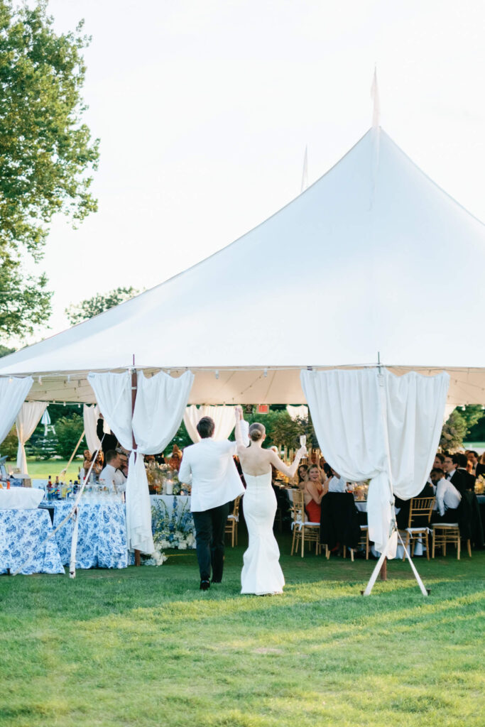 Bride and groom making their joyful grand entrance into a sailcloth reception tent, greeted by applauding guests and floral-draped décor.