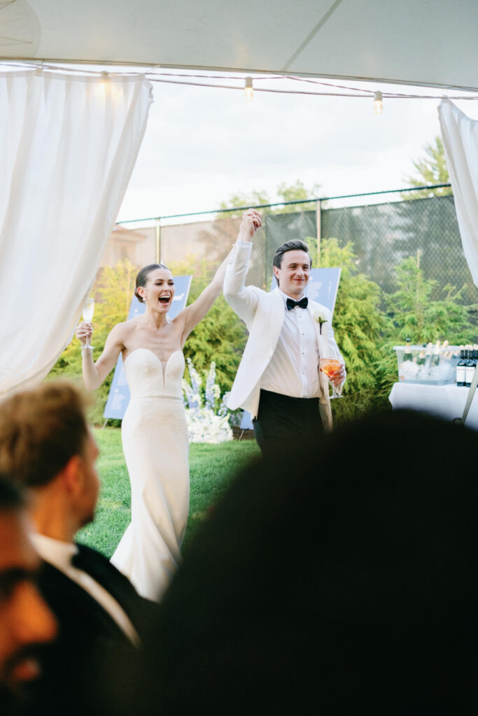 Bride and groom raise their joined hands in celebration as they enter their tented reception, grinning while holding champagne and Aperol spritzes.