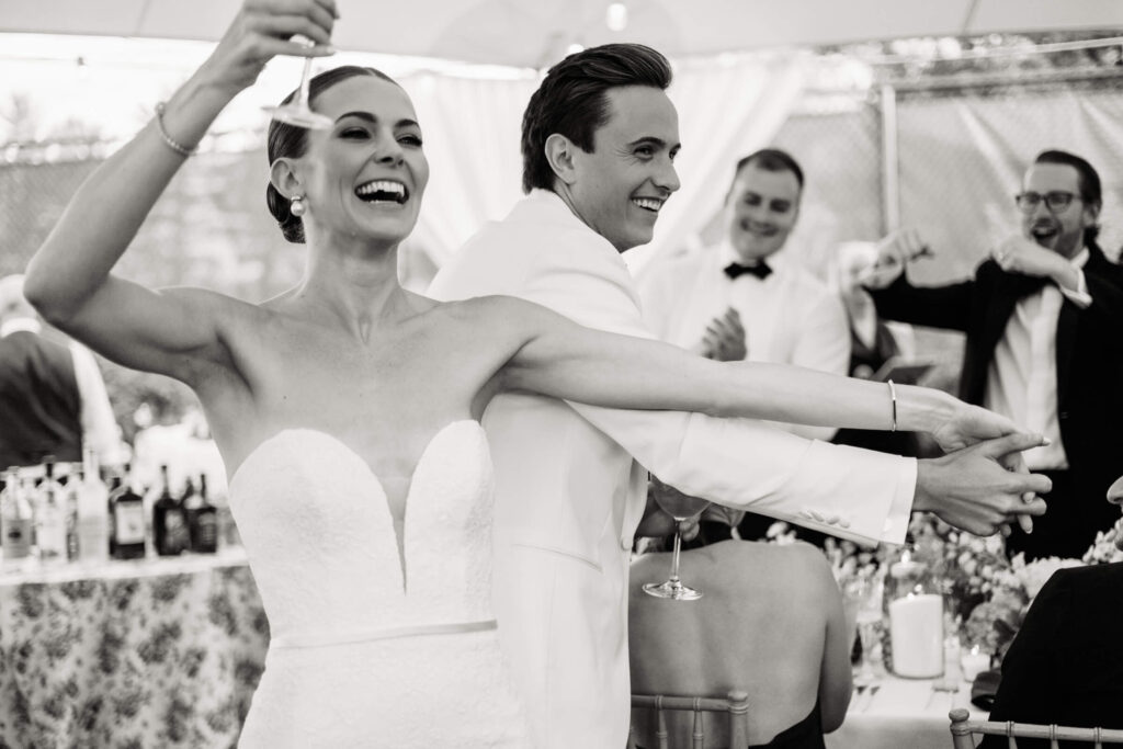 Joyful moment of the bride raising her glass in a toast, laughing with guests during the wedding reception under twinkle lights.