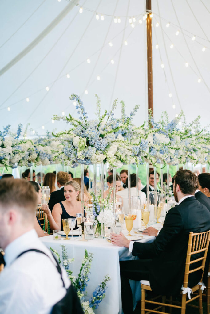Guests seated under the sailcloth tent surrounded by towering floral centerpieces in white and dusty blue, sharing conversations during dinner.