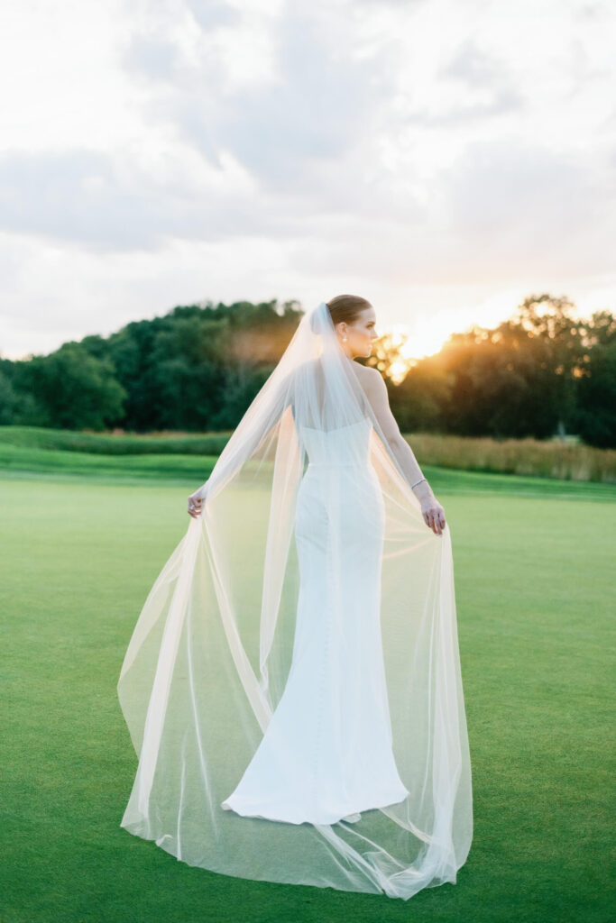 Bride stands alone in an open field during golden hour, her cathedral veil flowing in the breeze as the sun sets behind her.