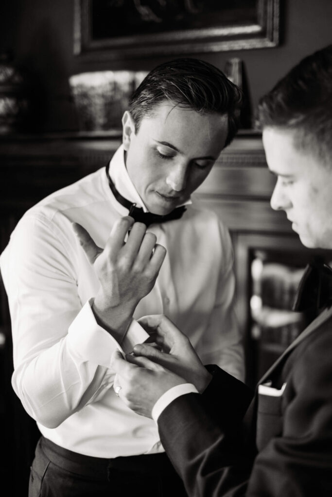 Black and white photo of the groom adjusting his cufflinks as his best man helps him get ready in the library at Castle Hotel & Spa