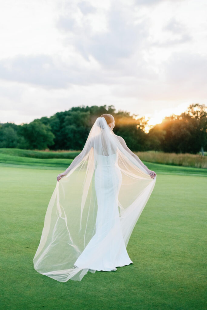 Bride walks across a golf course at sunset, her long veil trailing behind her in the golden light.