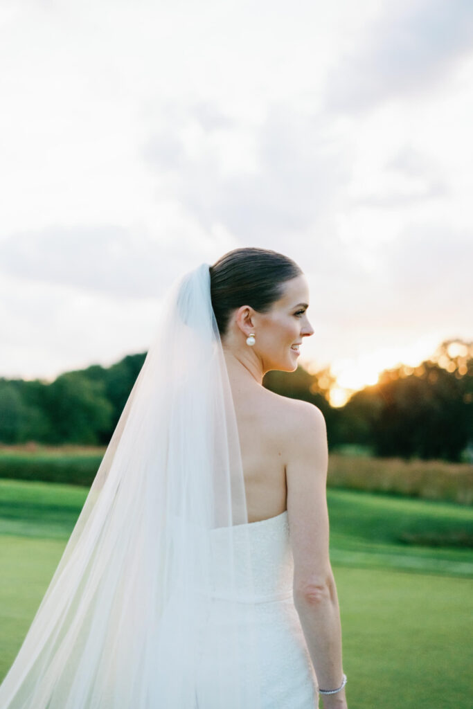 Close-up of bride looking over her shoulder, smiling softly with her veil cascading down her back as the sun sets.
