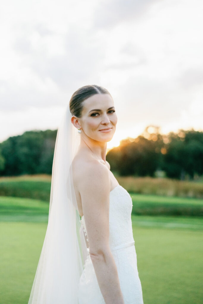 Elegant bride turns slightly toward the camera with a gentle smile, backlit by the soft glow of a summer sunset.