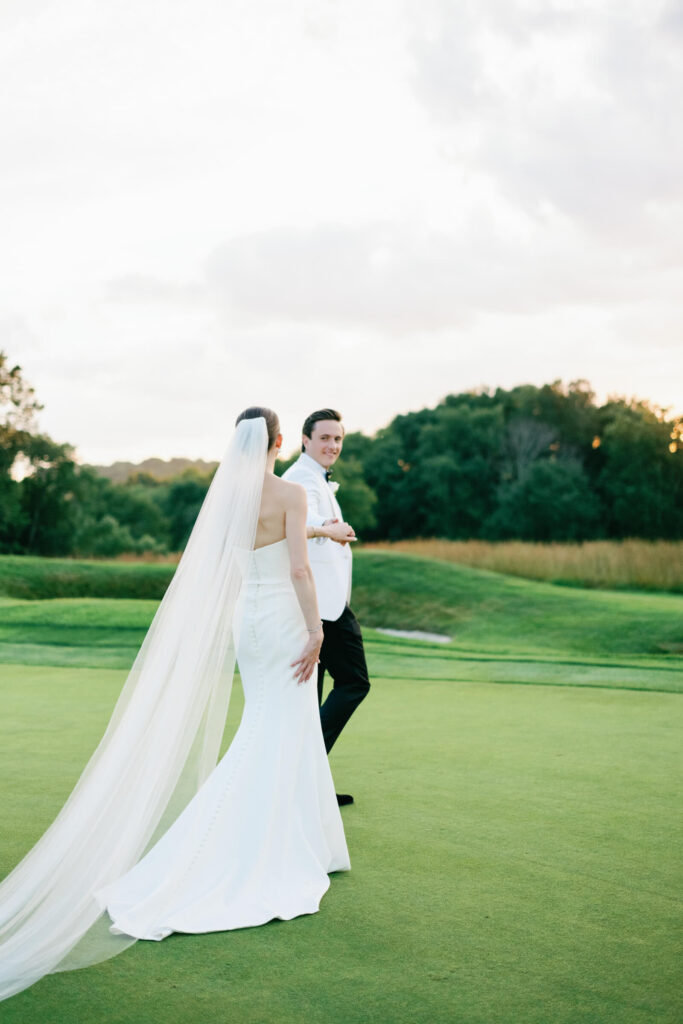 Groom holds bride’s hand as they walk together on the golf course, surrounded by rolling green hills.