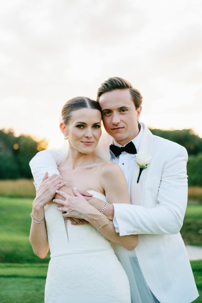Groom wraps his arms around the bride from behind as they pose confidently together at dusk.