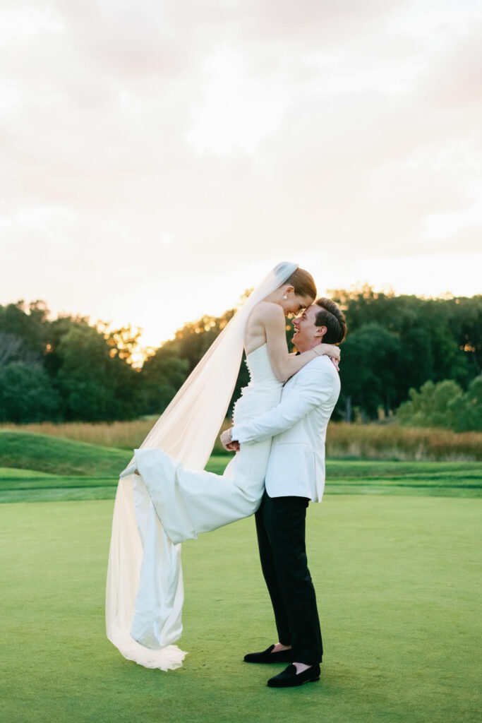 Groom lifts the bride in a joyful embrace as her veil flows behind them on the vibrant green lawn.