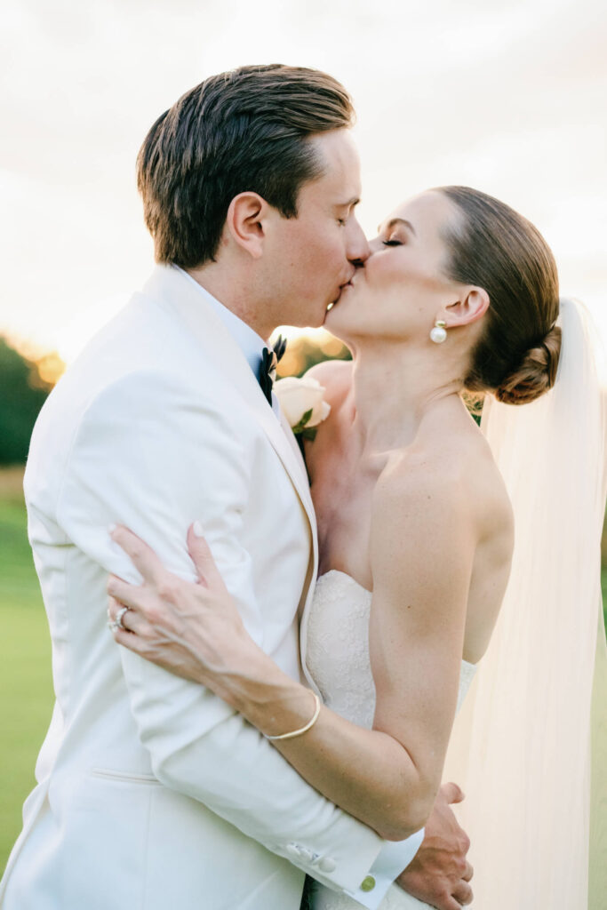 Bride and groom share a romantic kiss on the golf course, wrapped in each other’s arms as the sun dips behind the trees.