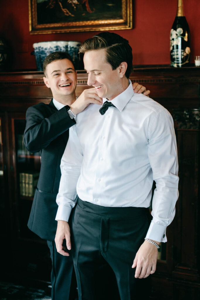 Groom smiling as his groomsman helps with his tie, captured in front of a warm wood-paneled fireplace at Castle Hotel & Spa