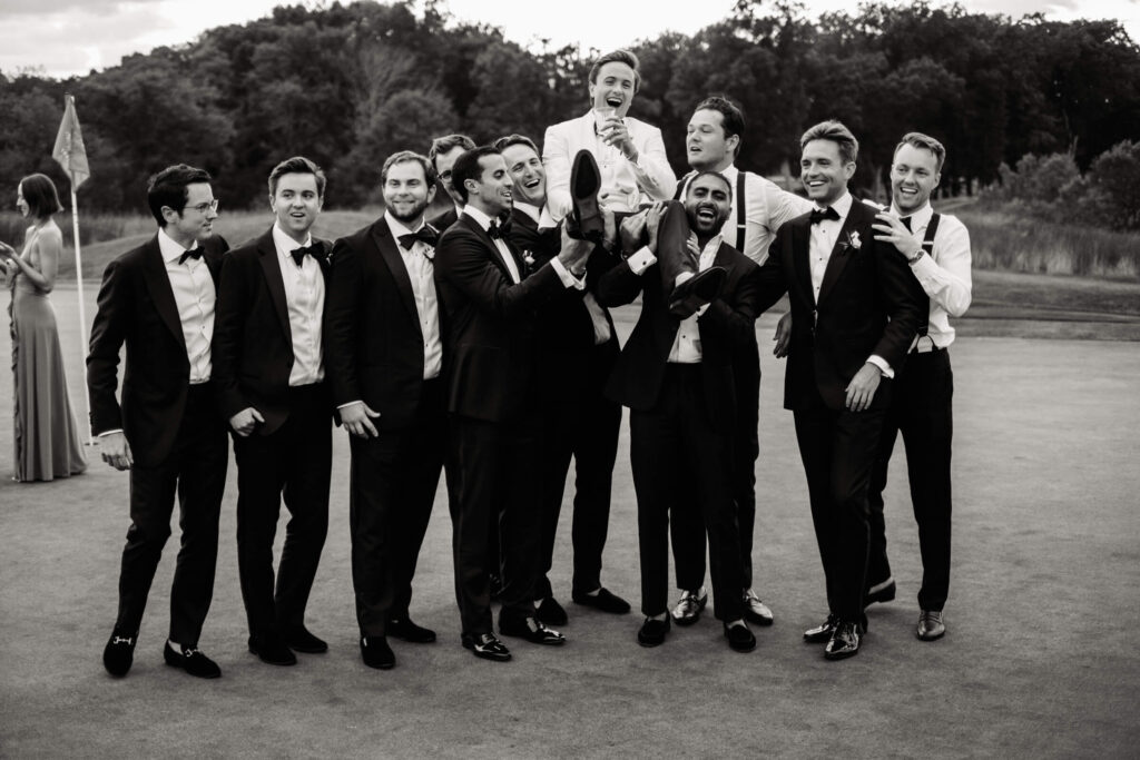 Groom being lifted in the air by groomsmen during celebratory group photo on the golf course, all dressed in black tie attire.
