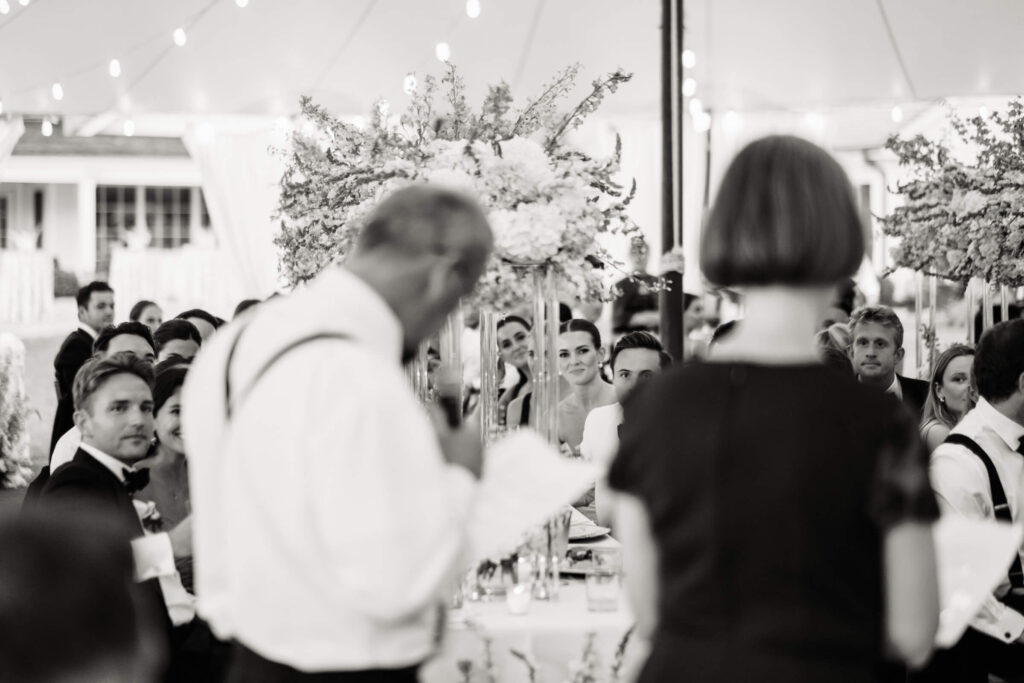 Bride and groom seated under a floral canopy, listening intently to a toast being given during the wedding reception.