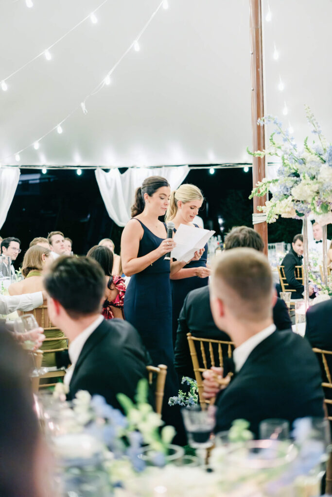 Two bridesmaids delivering a speech at the reception, standing under a tent strung with twinkle lights.