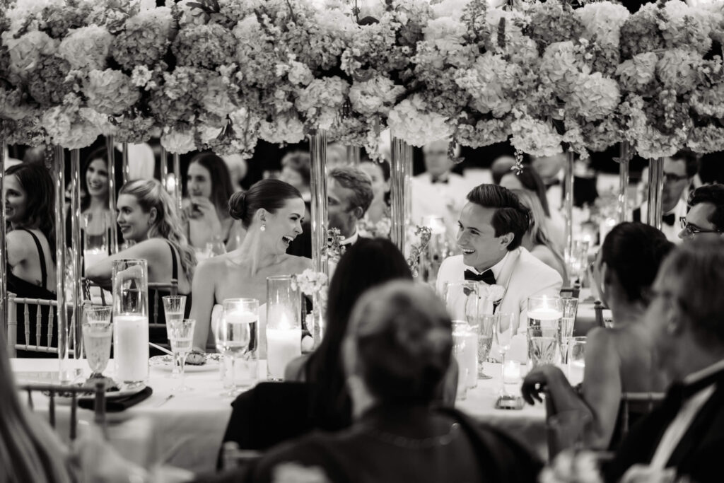 Bride and groom sharing a joyful moment at their candlelit head table beneath a lush floral installation.