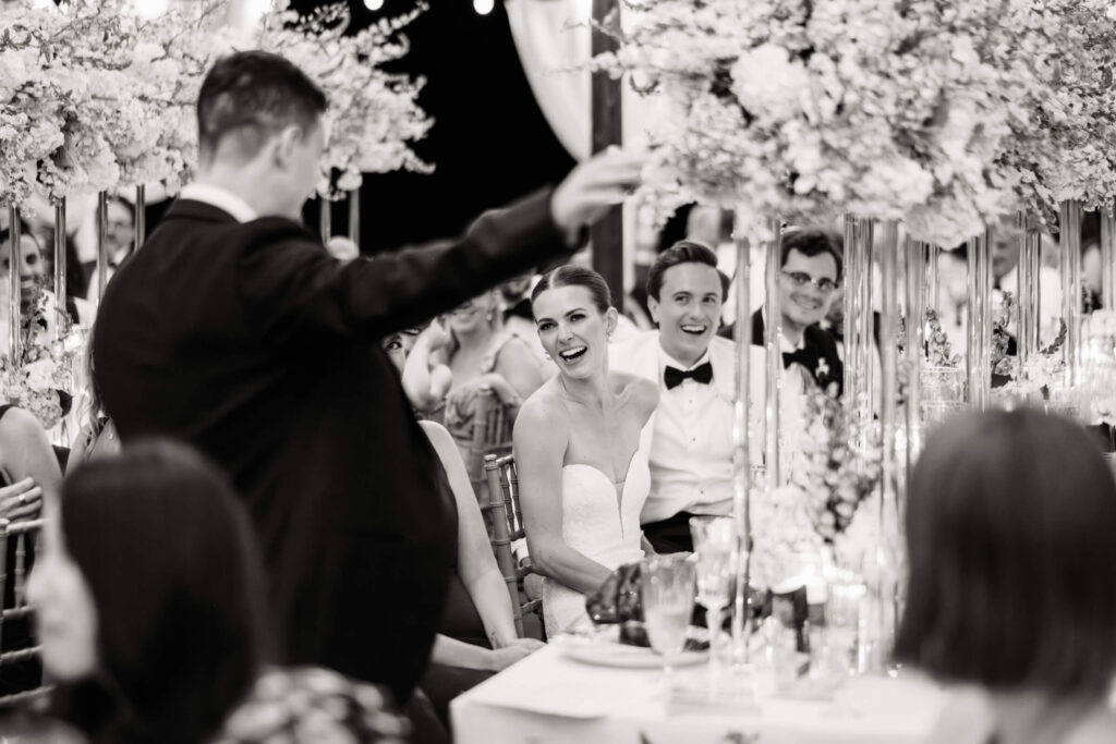 Bride and groom laughing as a guest gives a lively toast at their reception, surrounded by loved ones.