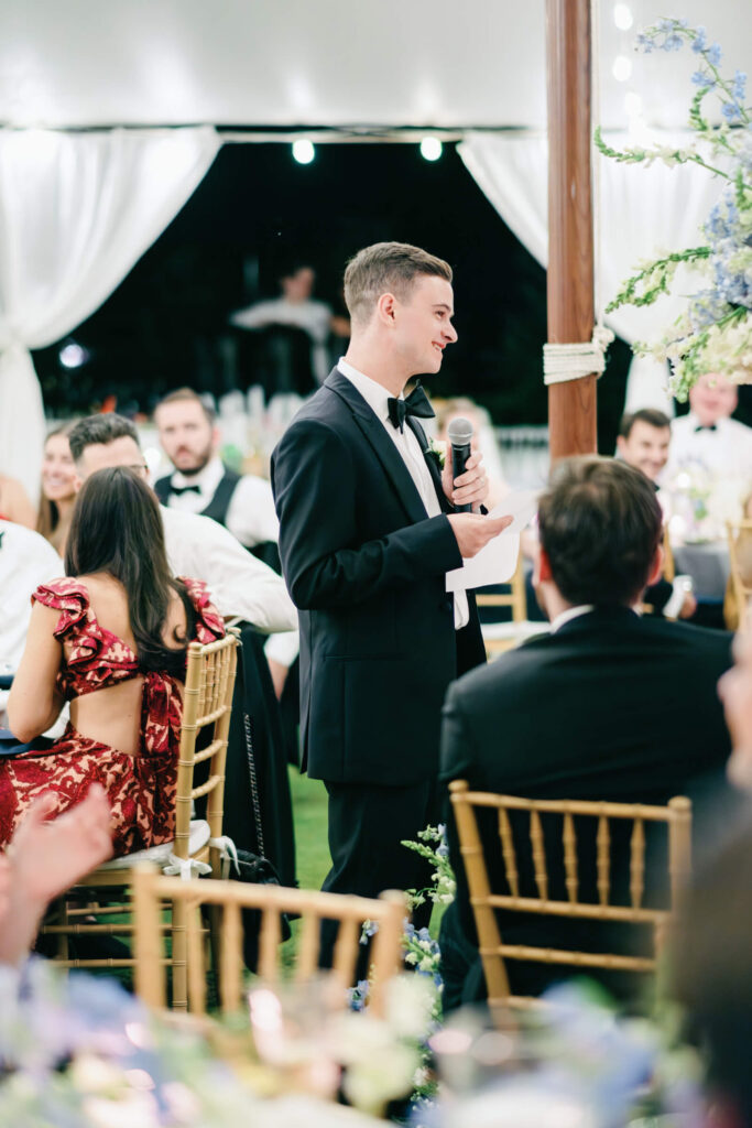Young man giving a heartfelt speech during the wedding reception, smiling while holding a microphone.