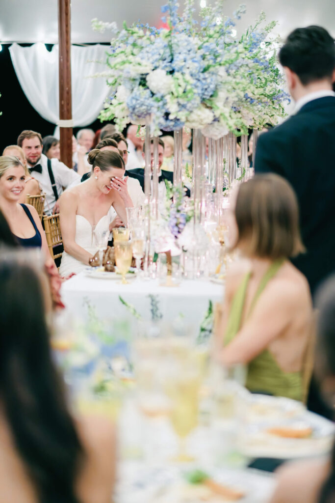 Bride covering her face with laughter during a toast, seated at a reception table with blue and white floral arrangements.