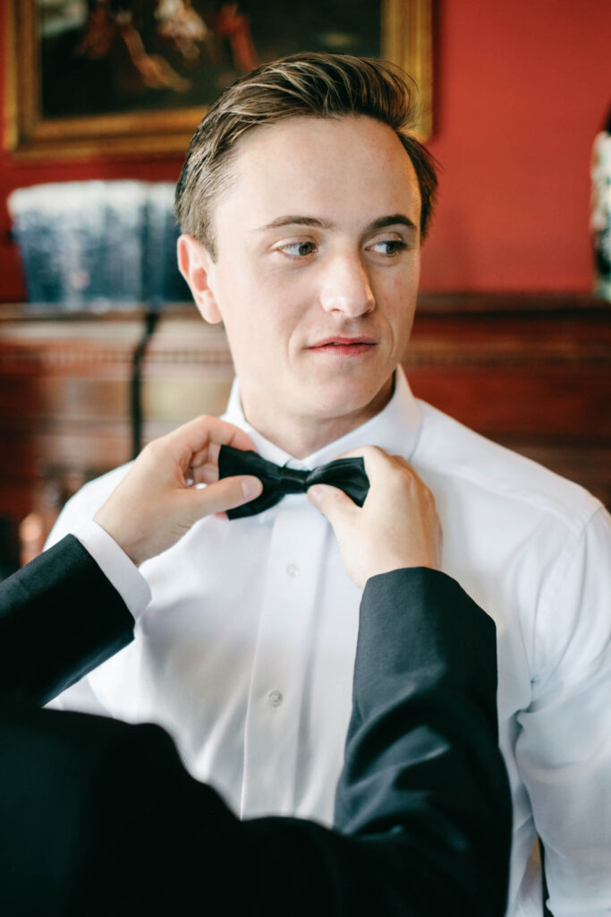 Close-up of the groom having his bowtie adjusted, framed against a deep red wall with vintage décor at the Castle Hotel & Spa