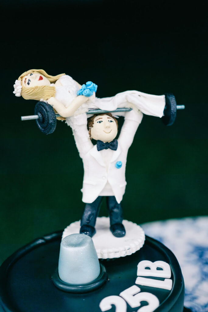 A playful cake topper featuring a groom in a tuxedo lifting the bride on a barbell, styled like a 25lb weight plate.