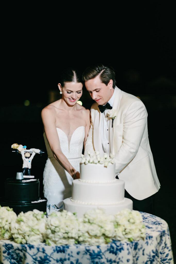 Bride and groom smile as they cut a classic white tiered cake, with a whimsical groom’s cake beside them.