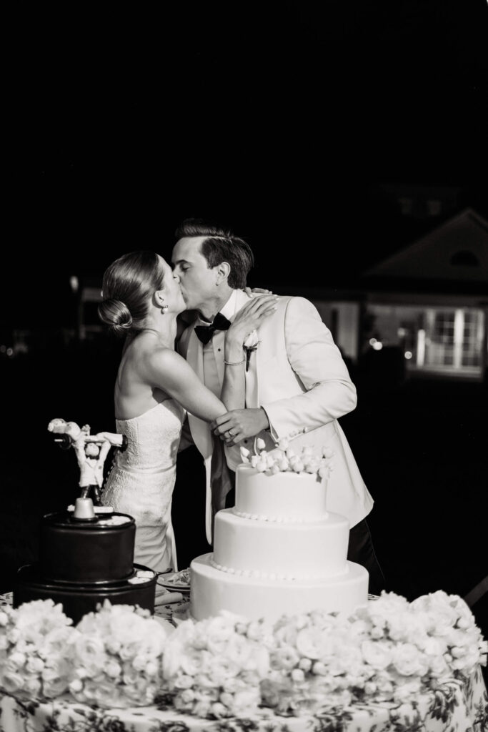 Bride and groom share a kiss after cutting their wedding cake, framed by soft floral arrangements and joyful ambiance.
