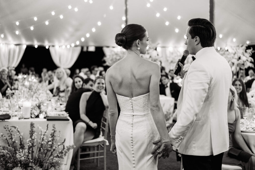 Bride and groom stand hand-in-hand under a canopy of string lights, addressing their guests with a heartfelt toast.