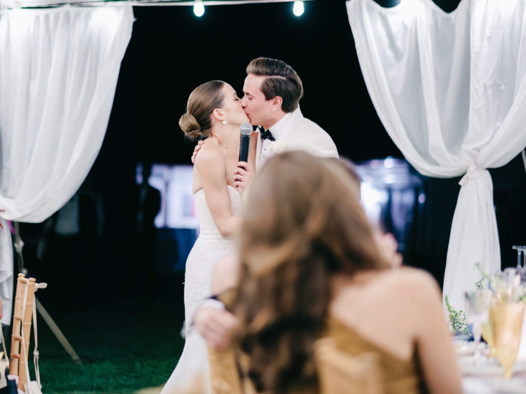Bride and groom exchange a kiss as they hold the microphone together, surrounded by draped white linens and glowing lights.