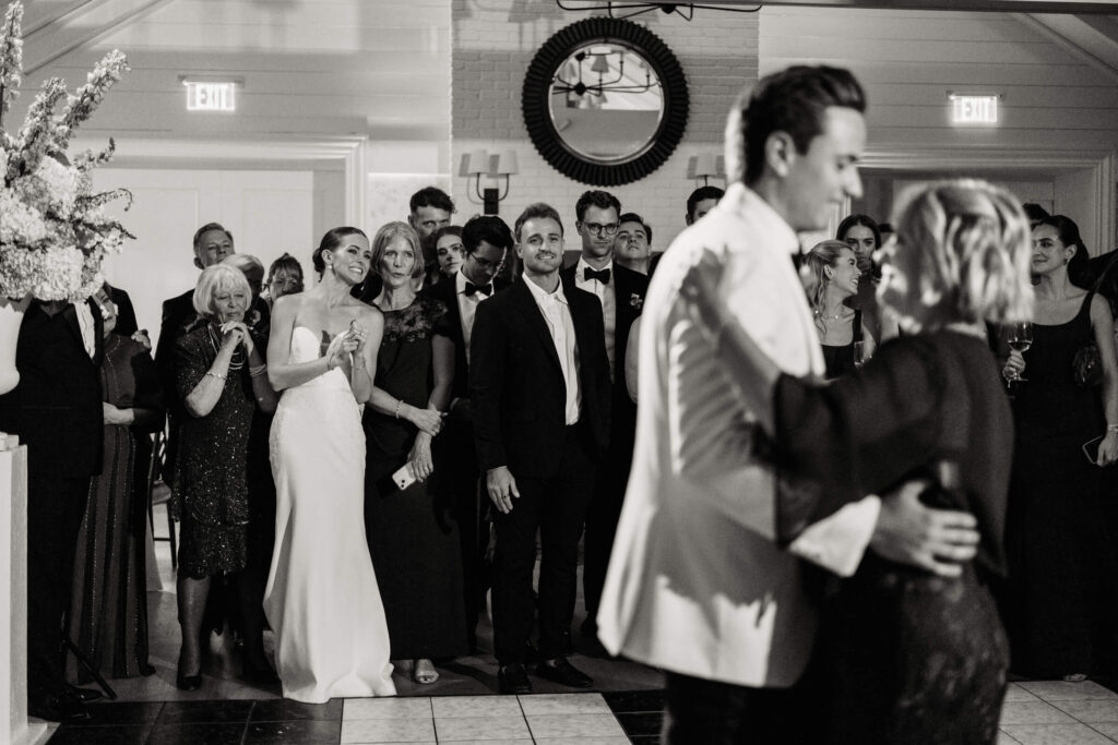 Bride beams with emotion as she watches her groom dance with his mother, flanked by close family and friends.