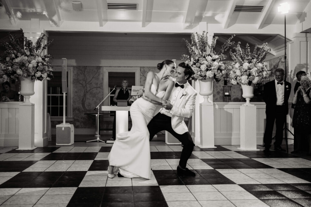 Groom dramatically dips his bride during their first dance, both laughing joyfully on a checkered dance floor.
