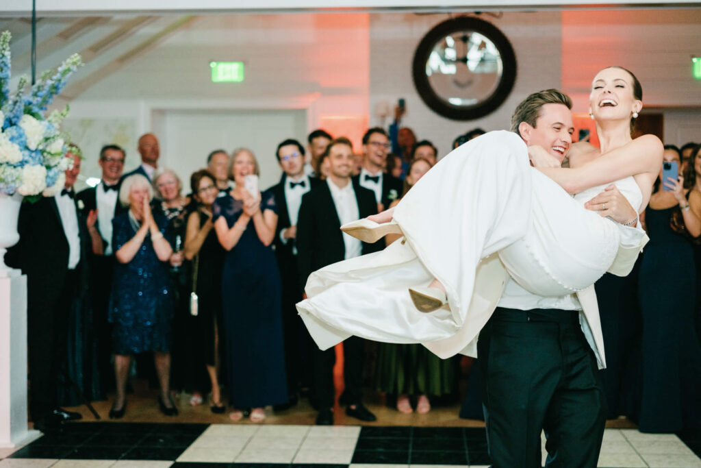 Groom lifts the bride into the air mid-dance, as guests cheer and smile in the background.