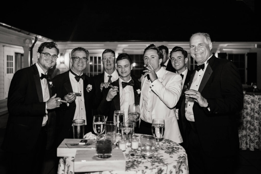 Groom and his groomsmen share a celebratory cigar moment with family, dressed in tuxedos and surrounded by glasses and floral linens.