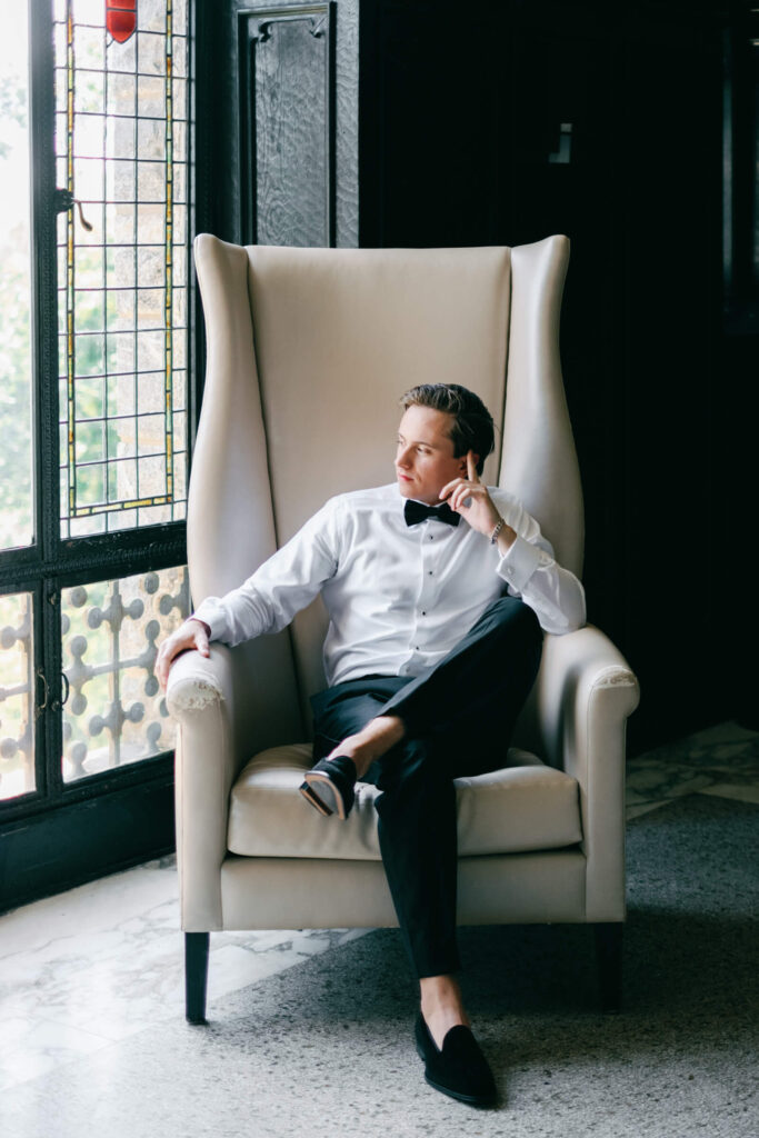 Groom seated in a high-back cream chair, gazing out the window in a moment of quiet before the New York country club wedding ceremony
