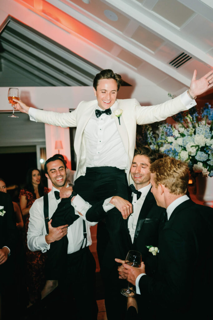 Groom being lifted in the air by friends during the reception, holding a wine glass with arms raised
