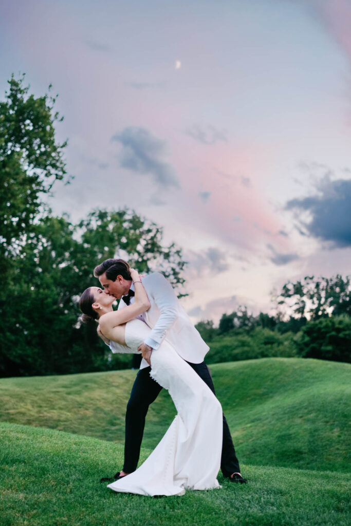 Bride and groom sharing a romantic kiss outdoors under a pastel twilight sky