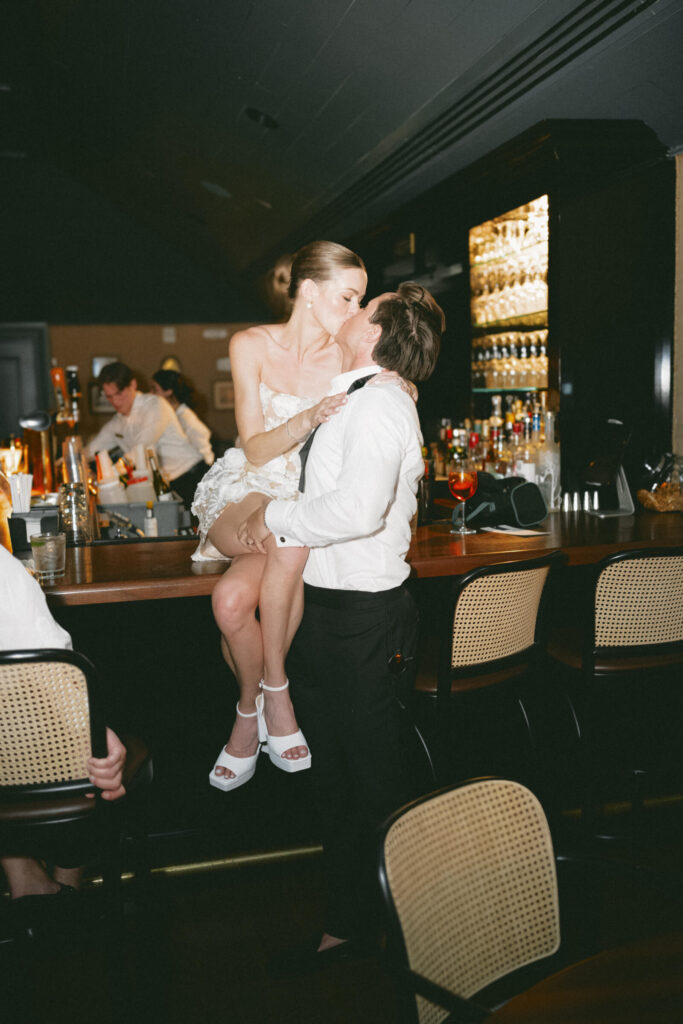 Groom lifting the bride onto the bar for a kiss during the wedding after-party