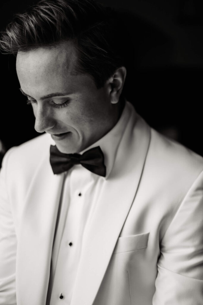 Black and white close-up of the groom wearing a white dinner jacket and bowtie, smiling softly as he prepares for the ceremony