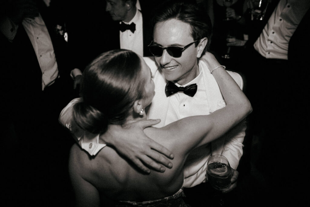 Bride and groom smiling and dancing closely together on the dance floor, surrounded by guests in formalwear.