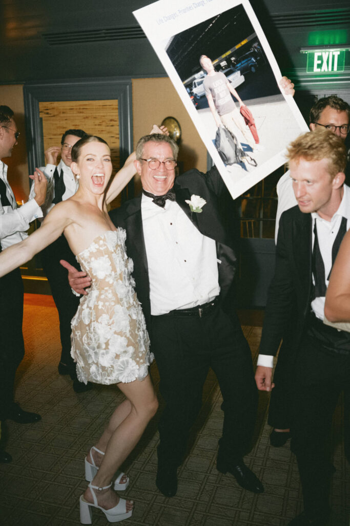 Bride and father of the bride laughing and dancing together, holding up a large humorous photo sign.