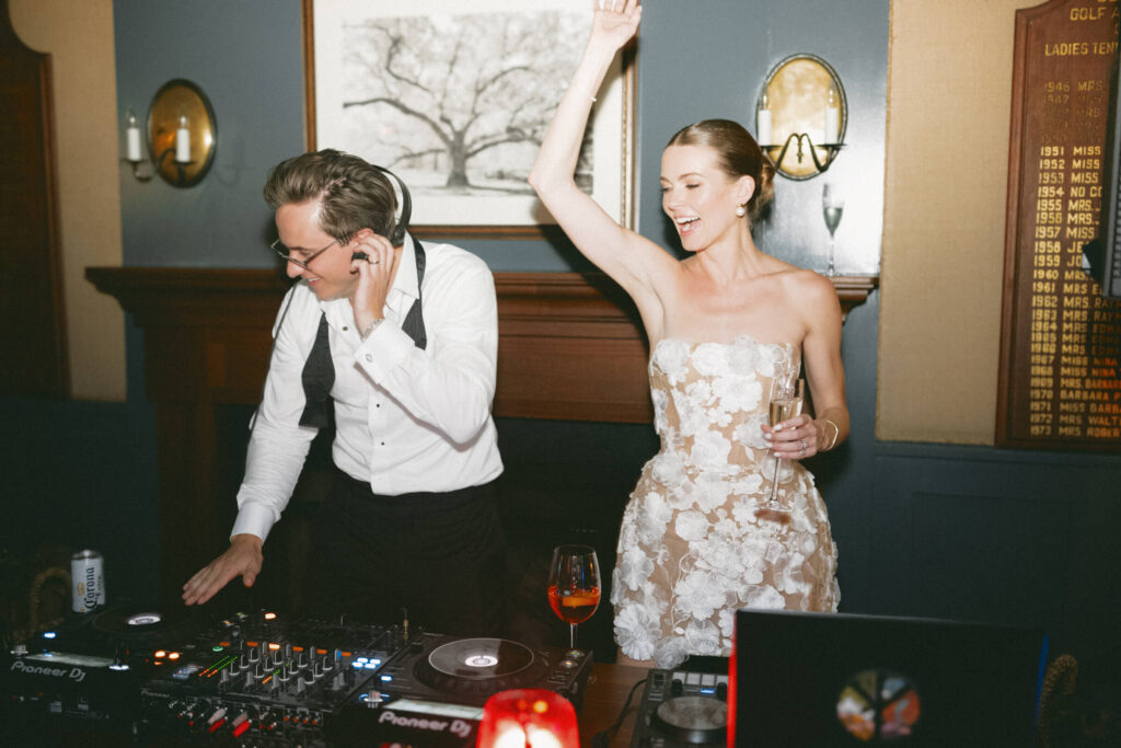 Bride and groom DJing together at the after party, both smiling and celebrating with champagne in hand.