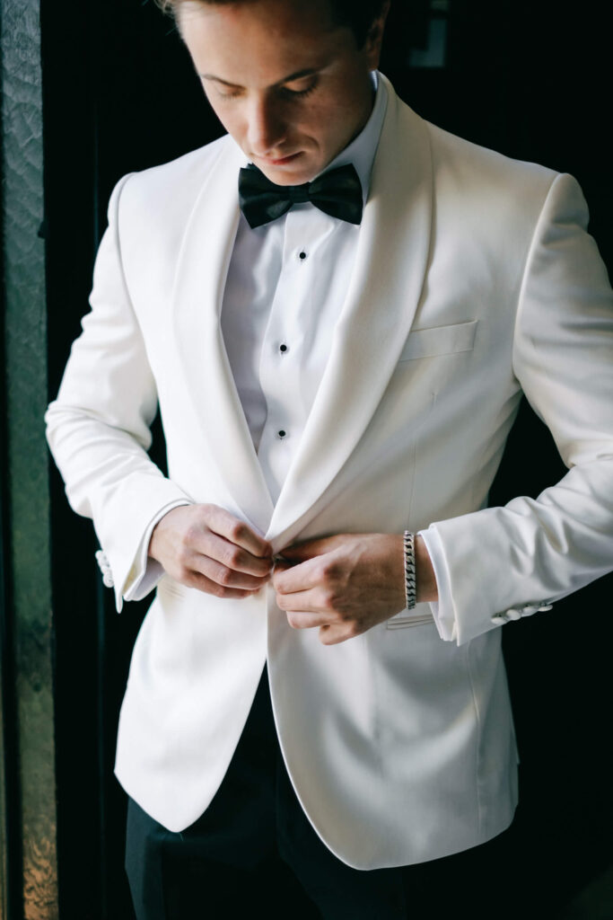 Groom adjusting the button of his white tuxedo jacket, showcasing his polished black bowtie and silver bracelet