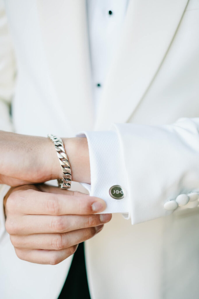 Detail shot of the groom’s monogrammed cufflink and silver bracelet against the sleeve of his white tuxedo jacket