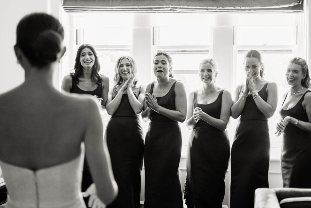 Bridesmaids reacting with joy and emotion as they see the bride in her gown for the first time, captured in black and white